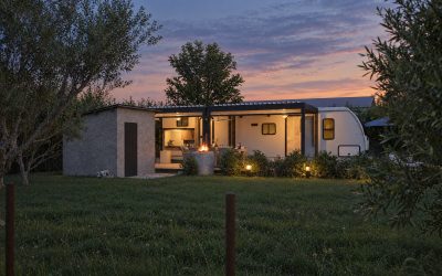 Camper, pergola and utility shed lit from within at blue hour across the lawn, fire pit burning